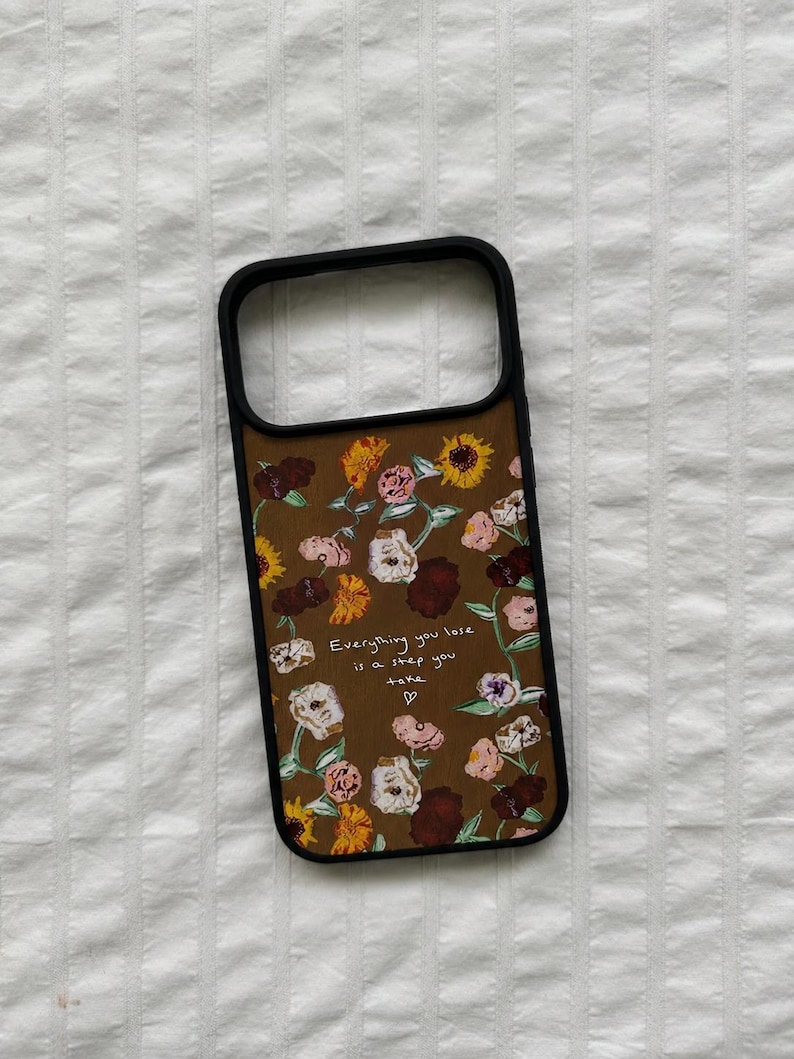 May include: A brown phone case with a floral pattern featuring sunflowers, roses, and other flowers. The case has a black border and the text "Everything you lose is a step you take" in white.