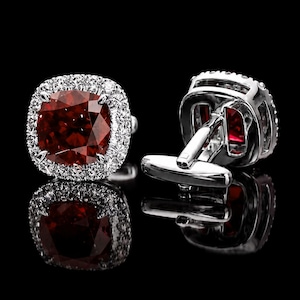 Puede incluir: Un par de gemelos plateados con una piedra preciosa de color rojo intenso de corte cuadrado en el centro, rodeada por un halo de pequeñas piedras transparentes. El marco de metal es pulido y reflectante, con un cierre de barra en T.