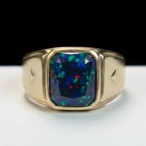 May include: A gold ring with a large, rectangular black opal gemstone. The opal displays a vibrant play of color with flashes of blue, green, and red. The ring has a classic, signet-style design with subtle triangular accents on the sides.