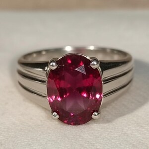 May include: A silver ring featuring a large, oval-cut, deep red gemstone. The ring has a simple band with two parallel grooves on each side of the gemstone setting. The gemstone is the focal point, showcasing its vibrant color and facets.