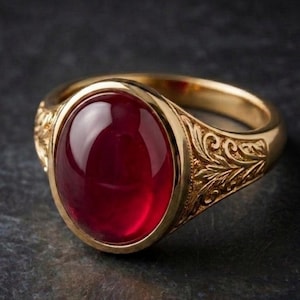 May include: A gold ring with an oval, deep red gemstone. The ring has an ornate, engraved design on the band. The gemstone is set in a gold bezel, and the ring is displayed against a dark background.