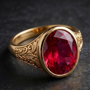May include: A gold ring with an oval, faceted, deep red gemstone. The ring has an ornate, engraved design on the band and around the gemstone setting. The ring is set against a dark background.