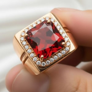 May include: A gold-toned ring featuring a large, square-cut, deep red gemstone. The gemstone is surrounded by a halo of small, clear gemstones. The ring has a thick band with a double-groove design, showcasing a classic, luxurious style.