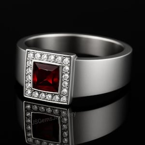 May include: A silver ring featuring a square, deep red gemstone, framed by smaller, clear stones. The ring has a wide band and a contemporary design. The image is set against a black background.