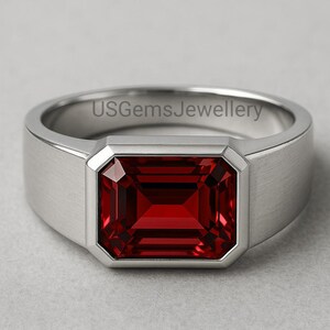 May include: A silver ring featuring a rectangular, deep red gemstone. The ring has a brushed metal finish and a modern design. The gemstone is set in a bezel setting, highlighting its rich colour.