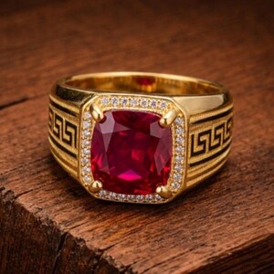 Könnte beinhalten: Ein goldener Ring mit einem großen, kissenförmigen roten Edelstein in der Mitte, umgeben von einem Kranz aus kleinen weißen Steinen. Die Ringschiene ist beidseitig mit einem schwarzen griechischen Mäandermuster verziert, das einen Kontrast bildet.