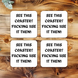 Op de afbeelding: Vier vierkante witte onderzetters met afgeronde hoeken, elk met de tekst "SEE THIS COASTER? FUCKING USE IT THEN!" in zwarte, vette letters. De onderzetters zijn gerangschikt op een houten oppervlak.