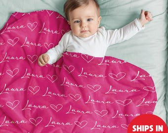May include: A hot pink baby blanket with the name "Laura" and heart shapes printed in white. The blanket is wrapped around a baby wearing a white onesie. The blanket is on a light blue bedsheet.