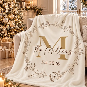 May include: A cream-colored fleece throw blanket featuring a large gold letter M, the text The Millers, and the text Est.2026. The design is encircled by a brown line-art floral wreath with delicate stems, leaves, and small flower buds.