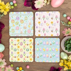 May include: Four square Easter-themed coasters with pastel designs. Each coaster features a different pattern of eggs, bunnies, chicks, and flowers in shades of blue, pink, yellow, and green. The coasters are arranged on a wooden surface with floral decorations.