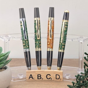 May include: Four ballpoint pens with circuit board designs. The pens have black and gold accents, with green and orange details. The pens are displayed in a clear acrylic pen holder. Scrabble tiles spell out the letters A, B, C, and D.