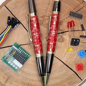 May include: Two ballpoint pens with red circuit board patterned barrels, gold accents, and black tips. The pens are displayed with electronic components, including LEDs, a circuit board, and a connector, on a wooden surface.