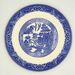 May include: A round, cream-colored plate with a blue Willow pattern design. The plate features a detailed border and a central scene with a pagoda, birds, and a boat. The plate is a classic piece of tableware.