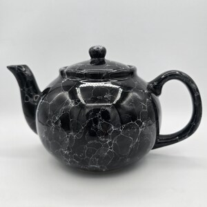 May include: A round ceramic teapot featuring a glossy black finish with a white marbled or bubble-like pattern throughout. The teapot includes a matching lid with a small knob, a curved spout on the left side, and a sturdy handle on the right side.
