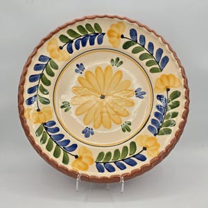May include: A round ceramic plate with a scalloped, reddish-brown rim. The cream-colored surface features a central yellow daisy-like flower surrounded by a border of painted yellow flowers, green leaves, and blue stems in a repeating pattern.