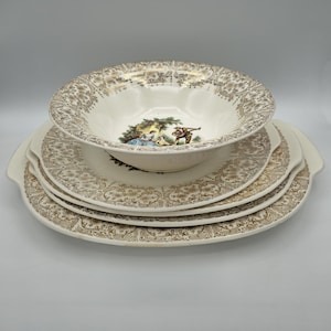 May include: A stack of vintage white and gold dinnerware. The set includes a large serving bowl and several plates with a gold filigree pattern. The bowl features a pastoral scene in the center. The plates have a scalloped edge.