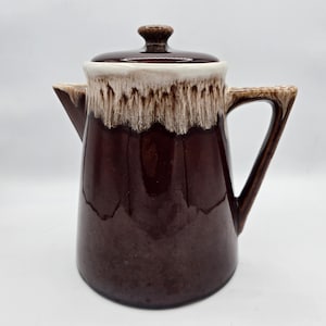 May include: A ceramic coffee pot with a glossy dark brown glaze and a contrasting cream-colored drip pattern around the top rim. The pot features a matching lid with a small knob handle, a side handle, and a spout, all finished in the same glaze.