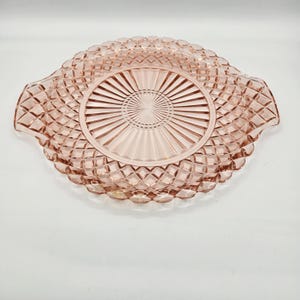 May include: A pink glass serving dish with a diamond pattern around the edges and a sunburst design in the center. The dish has two handles and is likely vintage.