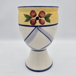 May include: A hand-painted ceramic goblet with a yellow rim and a floral design. The goblet features a cream-colored body with blue crisscross patterns and a pedestal base. The floral design includes maroon petals and green leaves.