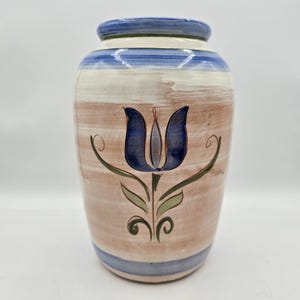 May include: A ceramic vase with a blue tulip design. The vase has a light brown base with white and blue horizontal stripes. The top and bottom rims are blue. The tulip is dark blue with green leaves and stems.