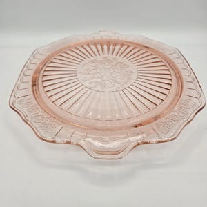 May include: A pink glass cake plate with a floral design in the center and along the scalloped edges. The plate has a ribbed pattern radiating from the center and two handles. A vintage-style serving dish.