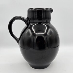 May include: A glossy black ceramic pitcher with a rounded, bulbous body and a curved handle. The vessel features a short, wide neck and a small, integrated pouring spout at the top rim, showcasing a smooth, reflective surface throughout the piece.
