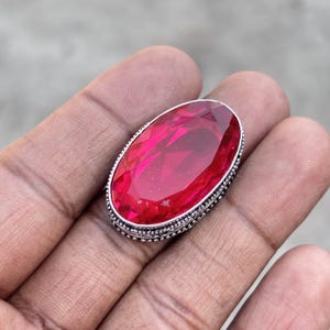 Puede incluir: Un anillo ovalado con una gran piedra preciosa roja facetada. El anillo tiene un borde plateado con cuentas. El anillo está sobre una mano.