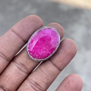 May include: A large, oval-shaped ring featuring a vibrant pink gemstone set in a silver-toned metal band. The gemstone has a textured surface and is surrounded by a beaded border. The ring is a statement piece.
