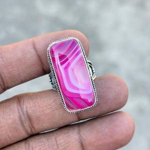 May include: A silver ring featuring a large, rectangular pink agate stone with white and pink banding. The stone is set in a decorative silver bezel with a detailed band.
