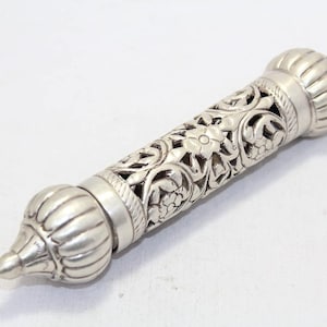May include: A silver-coloured, cylindrical object with intricate floral and leaf carvings. The ends feature bulbous, ribbed designs, and the centre has an openwork pattern. The design suggests a decorative or ornamental item.