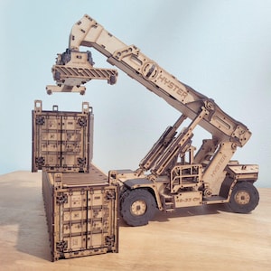 Puede incluir: Maqueta de madera de un manipulador de contenedores Hyster levantando un contenedor de envío. El modelo es de madera marrón claro y presenta la palabra "HYSTER" en el brazo y el cuerpo. Otros dos contenedores están en el suelo.