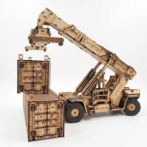 May include: A laser-cut wooden model of a Hyster reach stacker crane lifting a shipping container above another container. The model features intricate mechanical details, including wheels, a boom, and the Hyster brand name engraved on the side.