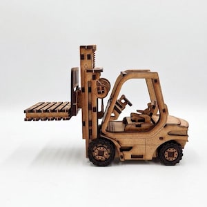 May include: A laser-cut wooden model of a forklift featuring a mast, a set of forks, a driver cabin with a seat and steering wheel, and four wheels. The light brown wood has dark, burnt edges from the cutting process, showing intricate assembly details.