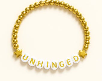 May include: Gold beaded bracelet with the word "UNHINGED" spelled out in white letter beads. Gold star-shaped beads flank the word. The bracelet is on a cream-colored background.