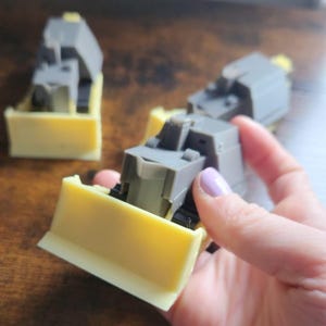 May include: A grey and yellow toy bulldozer. The toy features a grey cab and a yellow blade. The bulldozer is made of plastic and is designed to resemble a real construction vehicle. The toy is likely intended for children.