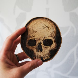 K&ouml;nnte beinhalten: Ein Untersetzer aus Holz mit einem schwarzen und wei&szlig;en Totenkopfdesign. Der Totenkopf ist in das Holz gebrannt und hat ein detailliertes Design.