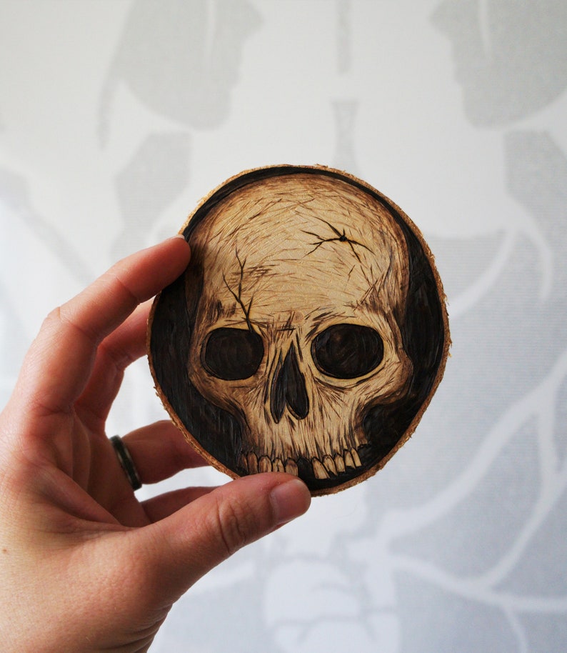 K&ouml;nnte beinhalten: Ein Untersetzer aus Holz mit einem schwarzen und wei&szlig;en Totenkopfdesign. Der Totenkopf ist in das Holz gebrannt und hat ein detailliertes Design.