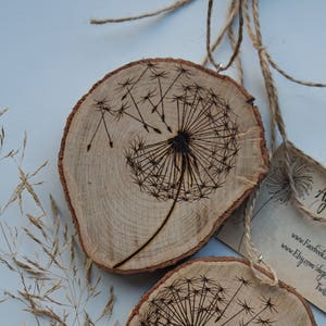 May include: Two wooden round slices with a woodburned dandelion design. The slices are hanging from twine and have a small tag attached with the text "Agni" and website information.