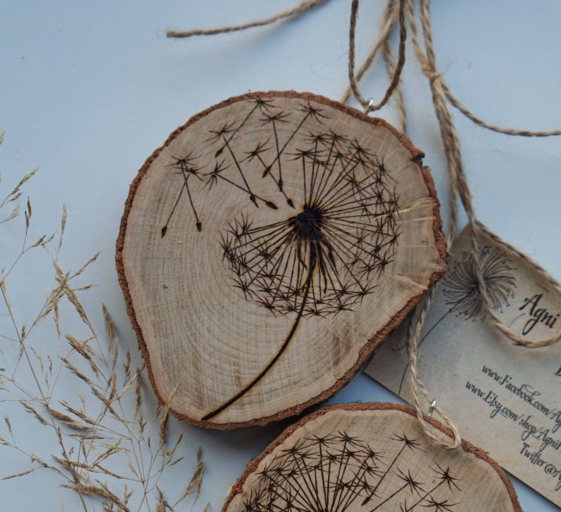 May include: Two wooden round slices with a woodburned dandelion design. The slices are hanging from twine and have a small tag attached with the text "Agni" and website information.