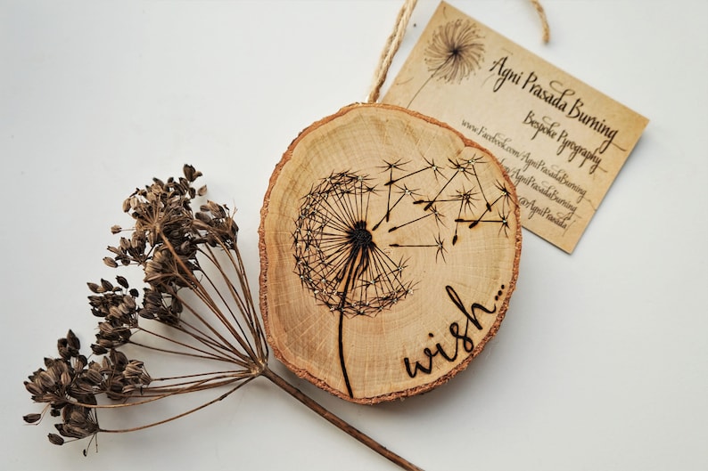 May include: A wooden slice with a burned design of a dandelion with the word "wish" written below it. The slice is hanging from a string and has a business card attached to it.