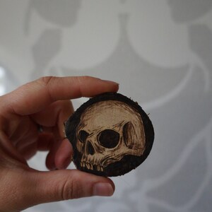 K&ouml;nnte beinhalten: Ein mit einem Brennstift auf ein rundes Holzscheibe gebranntes Totenkopfdesign. Der Totenkopf ist schwarz-wei&szlig; und hat ein detailliertes Design.