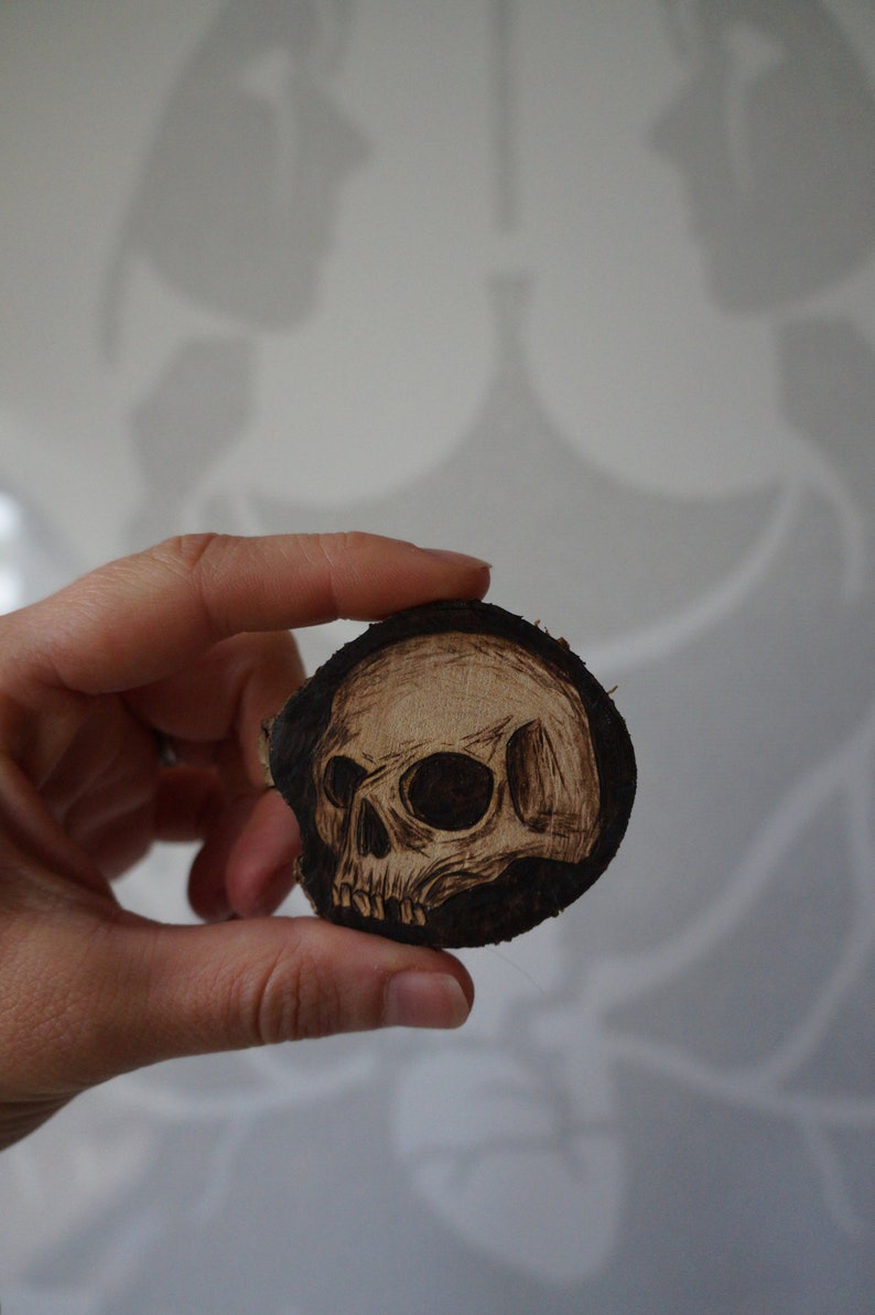 K&ouml;nnte beinhalten: Ein mit einem Brennstift auf ein rundes Holzscheibe gebranntes Totenkopfdesign. Der Totenkopf ist schwarz-wei&szlig; und hat ein detailliertes Design.