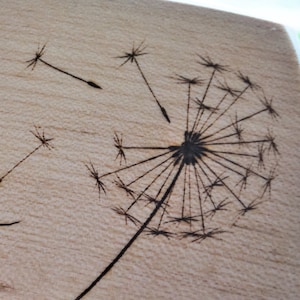 May include: A wood burned design of a dandelion with seeds blowing in the wind.