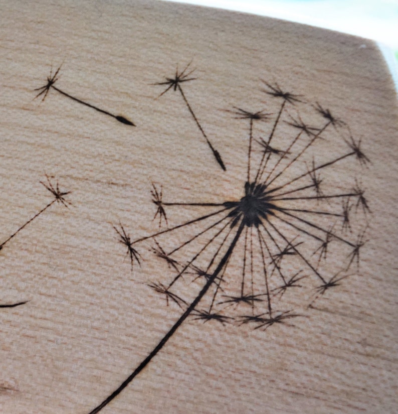 May include: A wood burned design of a dandelion with seeds blowing in the wind.
