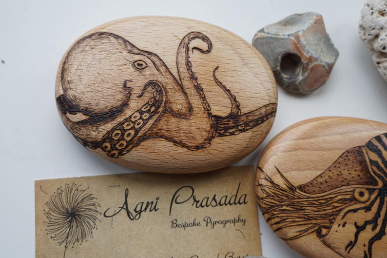 May include: A wooden plaque with a detailed woodburned image of an octopus with tentacles. The plaque is oval shaped and has a light brown color.