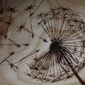 May include: A close-up of a wooden bowl with a burned-in design of a dandelion. The dandelion is in the centre of the bowl and has a dark brown colour. The dandelion has many seeds that are spread out around the bowl.