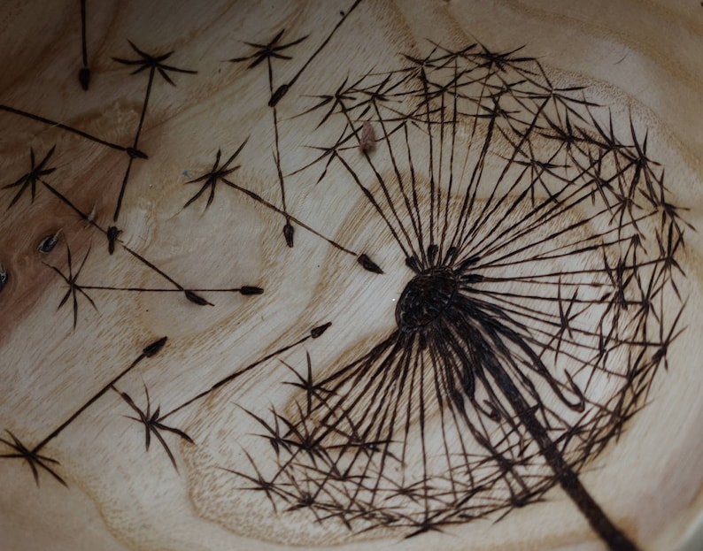 May include: A close-up of a wooden bowl with a burned-in design of a dandelion. The dandelion is in the centre of the bowl and has a dark brown colour. The dandelion has many seeds that are spread out around the bowl.