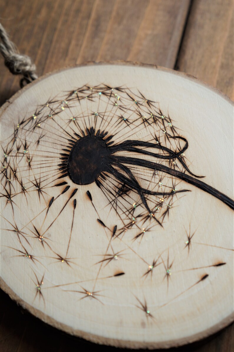 May include: A wood slice with a woodburned dandelion design. The dandelion is black and the seeds are scattered around the dandelion. The seeds have glitter on them.
