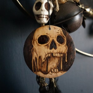 K&ouml;nnte beinhalten: Ein Holz-Anh&auml;nger mit einem Totenkopf-Design, der Totenkopf ist braun und schwarz mit tropfenden Details. Der Anh&auml;nger h&auml;ngt an einer Schnur mit einem wei&szlig;en Totenkopf-Anh&auml;nger und schwarzen Federn.