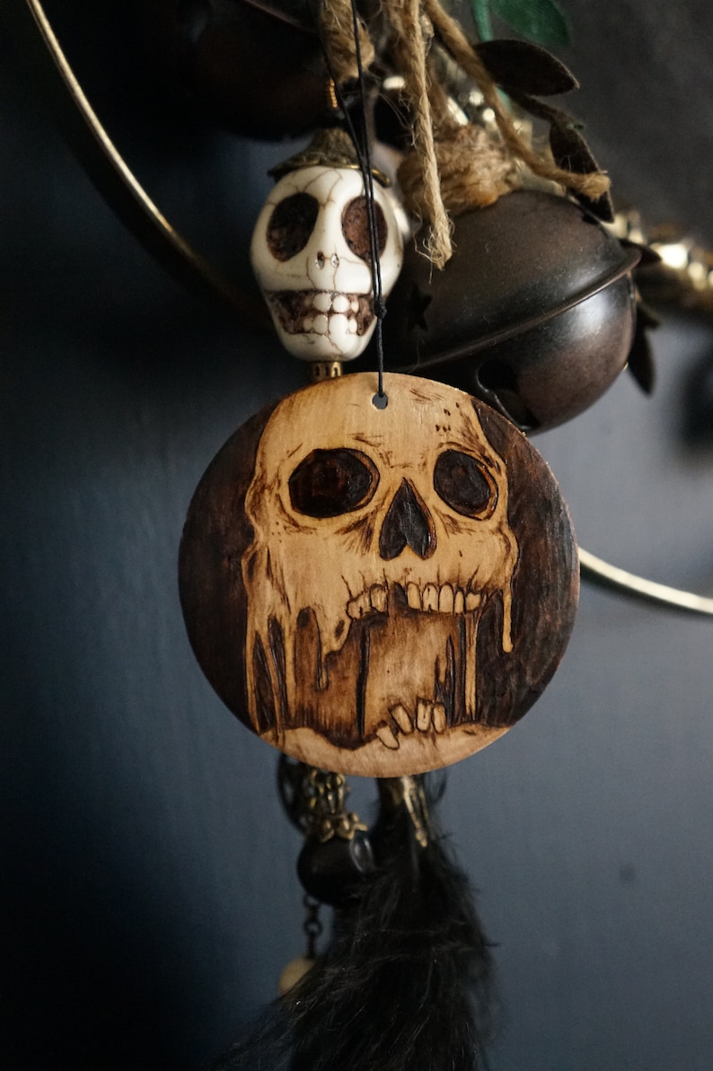 K&ouml;nnte beinhalten: Ein Holz-Anh&auml;nger mit einem Totenkopf-Design, der Totenkopf ist braun und schwarz mit tropfenden Details. Der Anh&auml;nger h&auml;ngt an einer Schnur mit einem wei&szlig;en Totenkopf-Anh&auml;nger und schwarzen Federn.